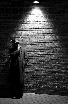 a man in a coat and hat standing next to a brick wall with a light on