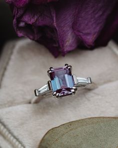 an engagement ring with two baguettes in front of a purple flower on a white box