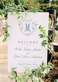 a welcome sign is surrounded by greenery in front of a building with people walking on the sidewalk