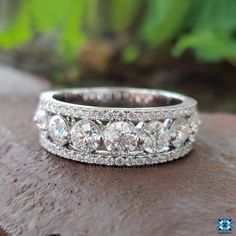 a wedding ring with three rows of diamonds on top of a wooden surface in front of green plants