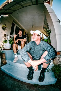 two men are sitting on the steps in front of their house and one is wearing a hat