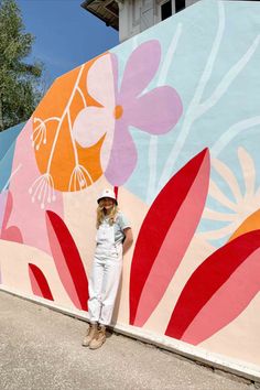 a woman standing in front of a colorful wall with flowers painted on it and wearing a hat