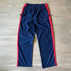 This Fresh Pair Of Nike Solid Swoosh Stripe Pant Is Made From A Lightweight Cozy Knit Satin Jersey Fabric, Keeping You Warm While Looking Dope At All Times. The Midnight Navy Blue Pant Features A Contrasting Red Stripe On The Side, With Minor Usa Flag Above The Back Pocket (Which Is Closable With A Zipper). The Nike Solid Swoosh Stripe Pant Has Two Side Pockets To Store All You Smaller Belongings Like Phone Or Wallet. - Elastic Waistband With Drawcord - Zippered Front Pockets - 100% Polyester Nike Blue Relaxed Fit Sweatpants, Blue Wide Leg Sportswear Pants, Sportswear Wide Leg Blue Bottoms, Sporty Blue Pants With Straight Hem, Blue Wide Leg Sportswear Bottoms, Sporty Red Straight Leg Bottoms, Red Nike Sports Pants, Nike Red Sports Pants, Nike Wide Leg Pants With Pockets