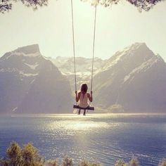 a woman sitting on a swing in the middle of a lake with mountains in the background