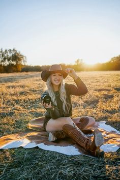 a woman sitting on top of a blanket in a field wearing boots and a cowboy hat