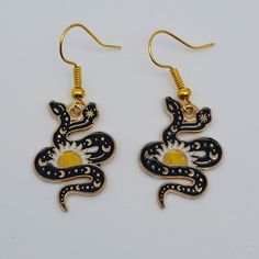 Add some charm and mystery to your look with these adorable cosmic serpent enamel earrings. Made from high-quality metal and glossy enamel, these earrings have hypoallergenic, nickel-free and lead-free gold-plated ear hooks. The snake is glossy black enamel with a celestial pattern of moon and stars on its body. Gold metal accents bring out every detail. The snake is wrapped around the sun, which is white with gold accents. The serpent is a symbol of rebirth, life, creation, and duality. The sun represents the source of life, energy, and light. The two heads of the snake represent the dual nature of life, such as male and female, light and dark, or order and chaos, or a symbol of the connection between the upper and lower worlds. Black Enamel Dangle Jewelry, Gold Enamel Earrings With Black Detail, Black Enamel Drop Earrings, Nickel-free Black Enamel Jewelry, Black Metal Snake-shaped Jewelry, Occult Earrings, Black Enamel Nickel-free Earrings, Spiritual Brass Snake-shaped Jewelry, Yellow Gold Snake-shaped Earrings