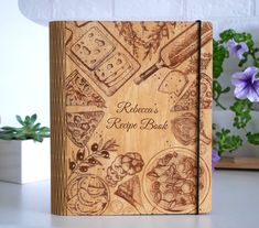 a wooden recipe book sitting on top of a table next to purple flowers and potted plants