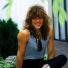 a man with long hair sitting on the ground in front of some bushes and plants