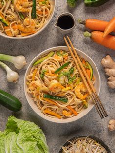 two bowls filled with noodles and vegetables next to carrots, broccoli, celery