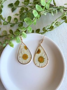 a pair of earrings with daisies in them on a white plate next to green leaves