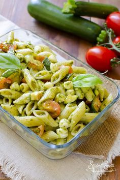 a glass dish filled with pasta and vegetables on a table next to cucumbers