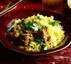 a red bowl filled with rice and meat next to a glass of beer on a table