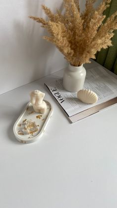 a white vase filled with dried flowers next to a book and seashell on a table