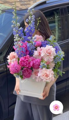 a woman holding a box full of flowers in front of a blue car with the door open