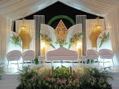 a stage set up with white chairs and floral arrangements on the side wall, surrounded by greenery