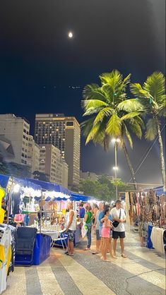 people are shopping at an outdoor market in the night time, with palm trees and buildings in the background