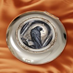 a silver plate with a painting of a black bird on it's side and an orange satin background