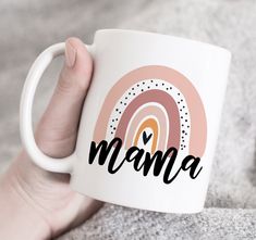 a person holding a white coffee mug with the word mama on it and a rainbow in the background