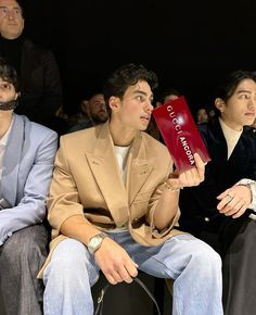 two men sitting next to each other at a fashion show, one holding a red box