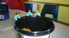 three lego people are standing in front of a trash can on the table next to some chairs