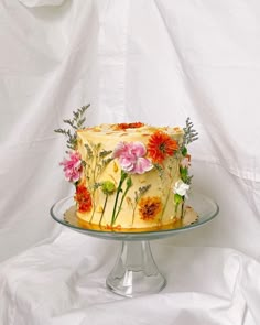 a yellow cake with flowers on it sitting on a glass stand against a white backdrop