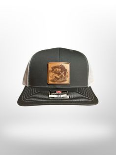 Elevate your style with the Richardson 112 Leather Patch Hat. This premium cap features a large mout bass design meticulously crafted on a genuine veg-tanned leather patch. The classic trucker style, combined with the durability of Richardson's high-quality materials, ensures both comfort and longevity. Perfect for outdoor enthusiasts and fashion-forward individuals alike, this hat offers an adjustable snapback closure for a custom fit. Whether you're by the water or in the city, make a bold sta Adjustable Flat Bill Snapback Hat For Fishing, Trucker Baseball Cap With Curved Brim For Fishing, Trucker Hat With Flat Bill For Fishing, Black Flat Bill Baseball Cap For Fishing, Brown Curved Brim Trucker Hat For Fishing, Trucker Hat For Fishing With Flat Bill, Trucker Snapback Hat With Flat Bill For Fishing, Brown Snapback Hat For Fishing, Brown Fishing Cap