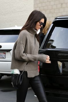 a woman in black leggings and a gray sweater is walking towards a car