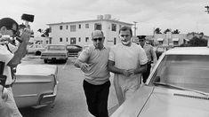 black and white photograph of two men walking in front of a car with another man talking on the phone