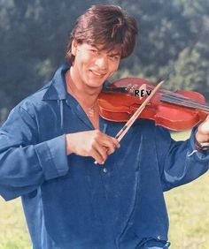 a man holding a violin in his right hand and smiling at the camera while wearing a blue shirt