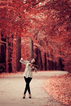 a woman is playing violin in the middle of an autumn tree - lined path with red leaves