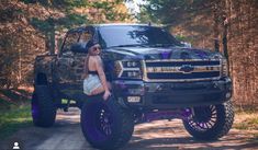 a woman sitting on the hood of a black truck in front of some tall trees