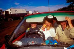 three women sitting in the driver's seat of a red car, one with a sticker on her head