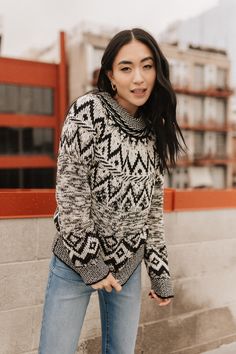 a woman standing in front of a wall wearing a sweater and jeans with her hands in her pockets