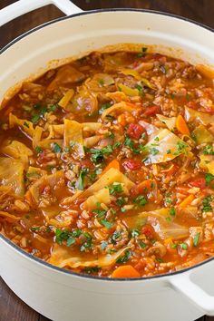 a white pot filled with vegetable soup on top of a wooden table next to a spoon