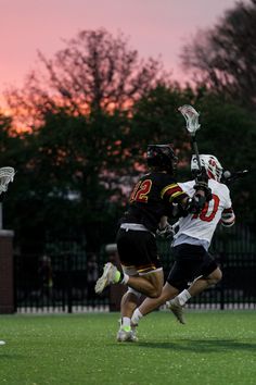 two men playing lacrosse against each other on a field at sunset with trees in the background