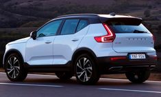 the rear end of a white volvo suv driving on a road with hills in the background