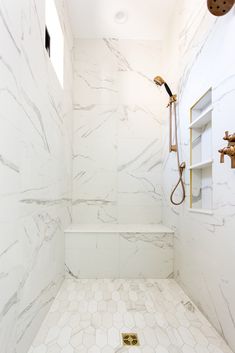 a white bathroom with hexagonal tiles and gold fixtures on the shower head, toilet, and shelf