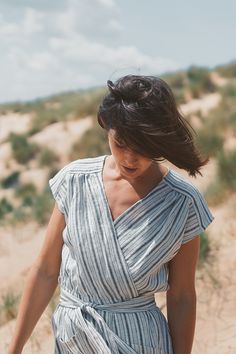 a woman in a striped dress walking on the beach with her hair blowing in the wind