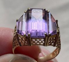 "14 karat yellow gold and amethyst ring.  The delicate filigree setting features three amethysts.  The center stone measures 9/16\" long and 5/8\" wide. It is an emerald cut amethyst flanked by 2 trapezoid cut amethysts.  The stones are of a light/medium violet color.  This is a statement ring in a simple symmetrical style.  Size is approximately 5." Art Deco Amethyst Ring In Yellow Gold, Yellow Gold Amethyst Ring In Art Deco Style, Purple Amethyst Ring With Intricate Design, Art Deco Yellow Gold Amethyst Ring, Purple Hallmarked Art Deco Amethyst Ring, Art Deco Purple Amethyst Ring, Formal Purple Amethyst Ring With Filigree, Purple Amethyst Ring Stamped 14k, Heirloom Amethyst Ring With Filigree