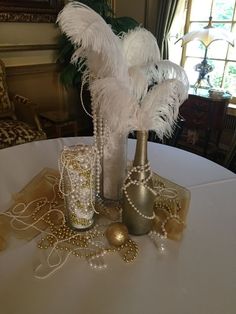 two vases with white feathers and beads on a table in front of a window