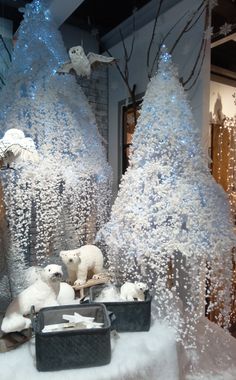 two white teddy bears sitting on top of a snow covered table next to fake trees