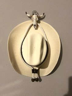 Ceramic Cattle Skull Cowboy Hat Hanger with Rope & Leather Accents Add a rustic, Western touch to your home with this unique cowboy hat hanger. Handcrafted with a ceramic cattle skull that exudes Southwestern charm, this display is perfect for showcasing your favorite cowboy hat. The natural white rope is paired with durable leather straps and rivets for a rugged yet elegant look. Finished with dark wooden beads at the ends, this piece blends classic cowboy aesthetics with modern craftsmanship. Whether you're a collector of Western décor or looking to add a stylish element to your space, this hanger is both functional and decorative. Perfect for entryways, living rooms, or bedrooms, it makes an ideal gift for Western enthusiasts or anyone wanting to bring a piece of cowboy culture into the Cowboy Hat Hanger, Skull Cowboy Hat, Cowboy Bedroom, Cattle Skull, Skull Cowboy, Cowboy Hut, Classic Cowboy, Hat Hanger, Chapeau Cowboy