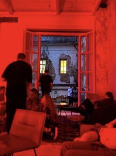 people sitting and standing in a room with red light coming through the window, while others stand around