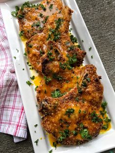 two pieces of meat covered in cheese and parsley on a white plate next to a red checkered napkin