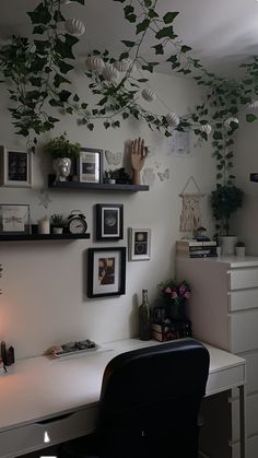 a white desk topped with a black chair next to a wall covered in pictures and plants