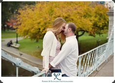 a man and woman kissing while standing on a bridge in front of a tree with yellow leaves