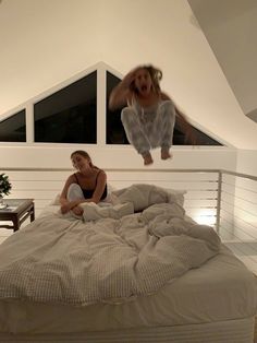 two women are sitting on a bed in a room with white walls and an open window