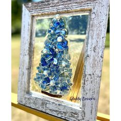 a blue glass christmas tree in a white frame on a wooden table with grass and trees in the background