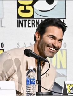 a smiling man sitting in front of a microphone at a press room table with water bottles