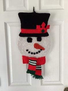 a crocheted snowman door hanger hanging on a white door with a black top hat and red scarf